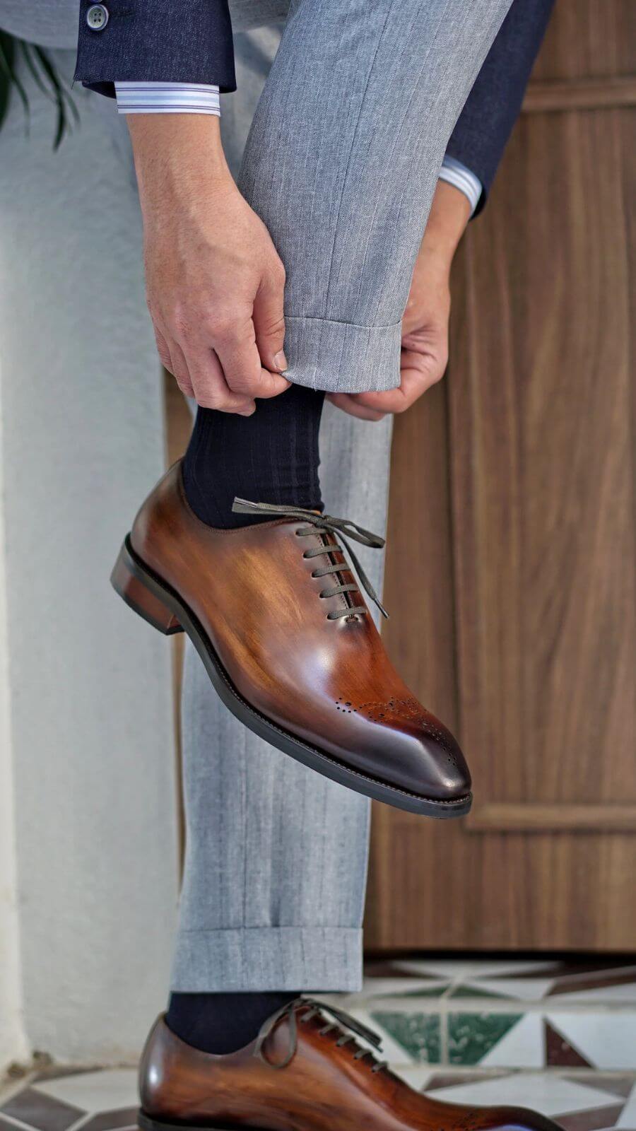 A pair of auburn wholecut oxford shoes with brogue detailing, displayed with wooden shoe trees on a wooden bench, leaning against a textured wall. The rich leather and elegant craftsmanship are highlighted by the natural lighting.