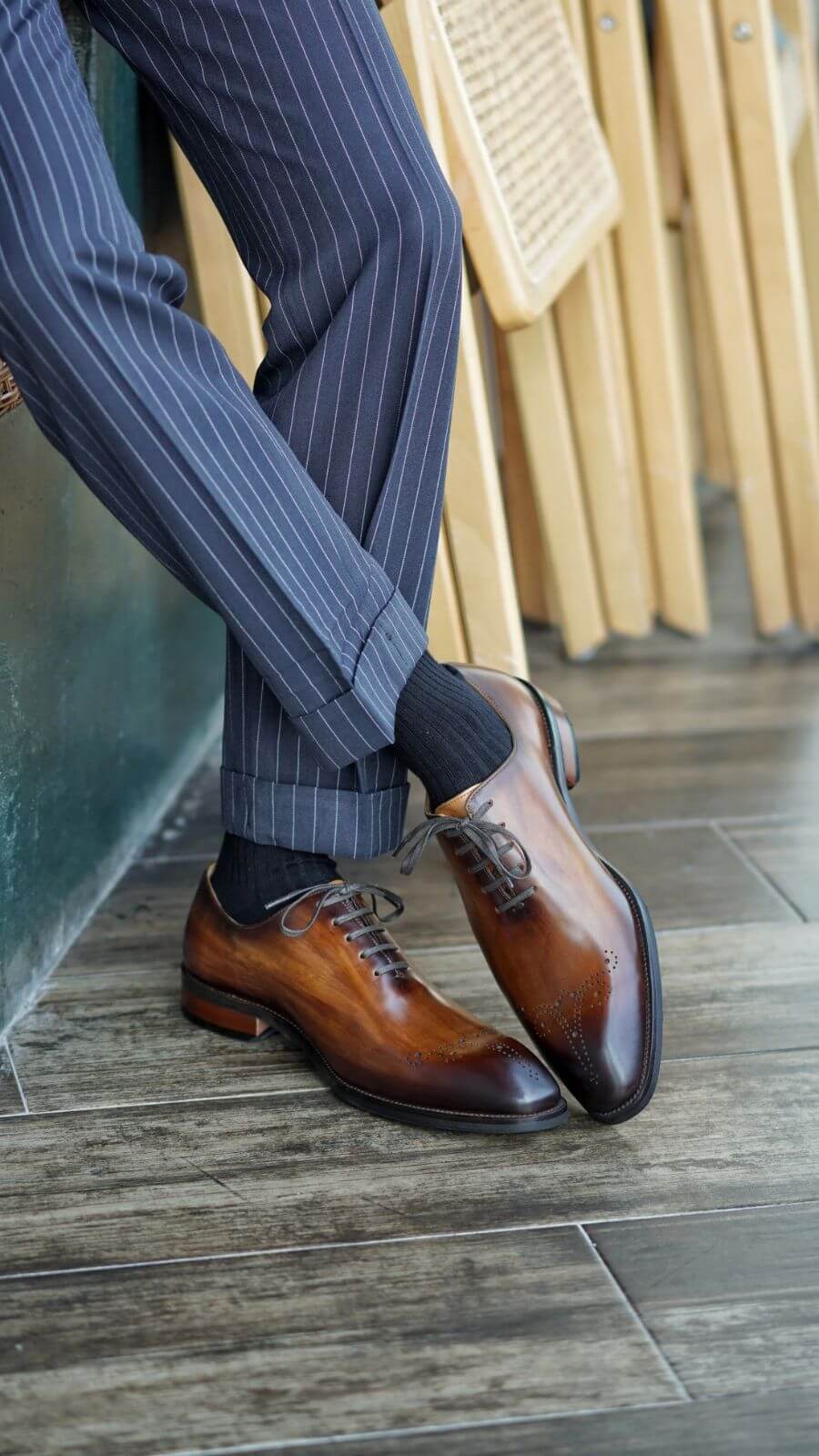 Close-up of a man wearing auburn wholecut oxford shoes paired with pinstriped navy trousers. The man is leaning against a wall with one leg crossed over the other, highlighting the elegant and polished leather shoes.