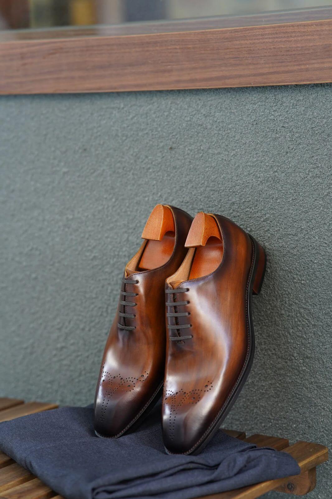 A pair of auburn wholecut oxford shoes with brogue detailing, displayed with wooden shoe trees on a wooden bench, leaning against a textured wall. The rich leather and elegant craftsmanship are highlighted by the natural lighting.