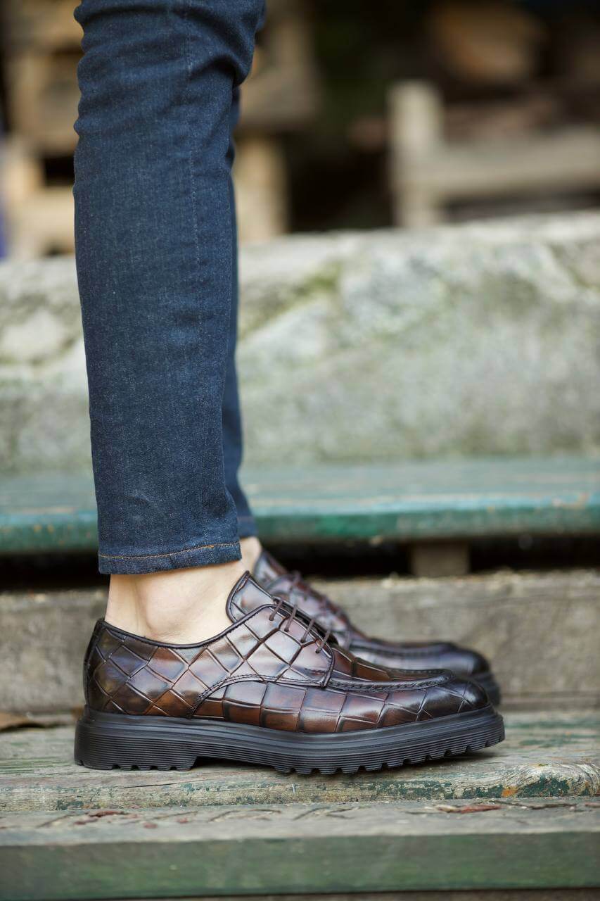 Close-up of a person wearing brown leather dress shoes with a textured pattern, stepping down on an old wooden staircase while dressed in dark denim jeans.