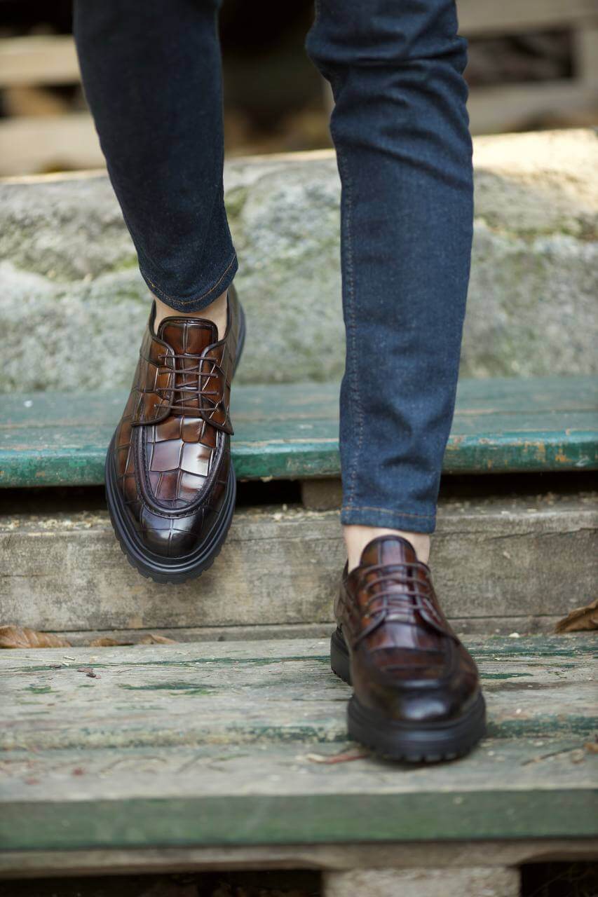 Close-up of a person wearing brown leather dress shoes with a textured pattern, stepping down on an old wooden staircase while dressed in dark denim jeans.