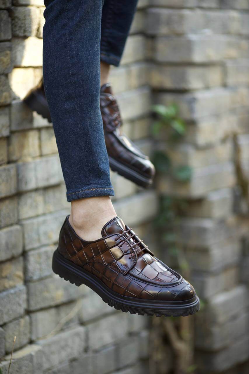 Close-up of a person wearing brown leather dress shoes with a textured pattern, stepping down on an old wooden staircase while dressed in dark denim jeans.