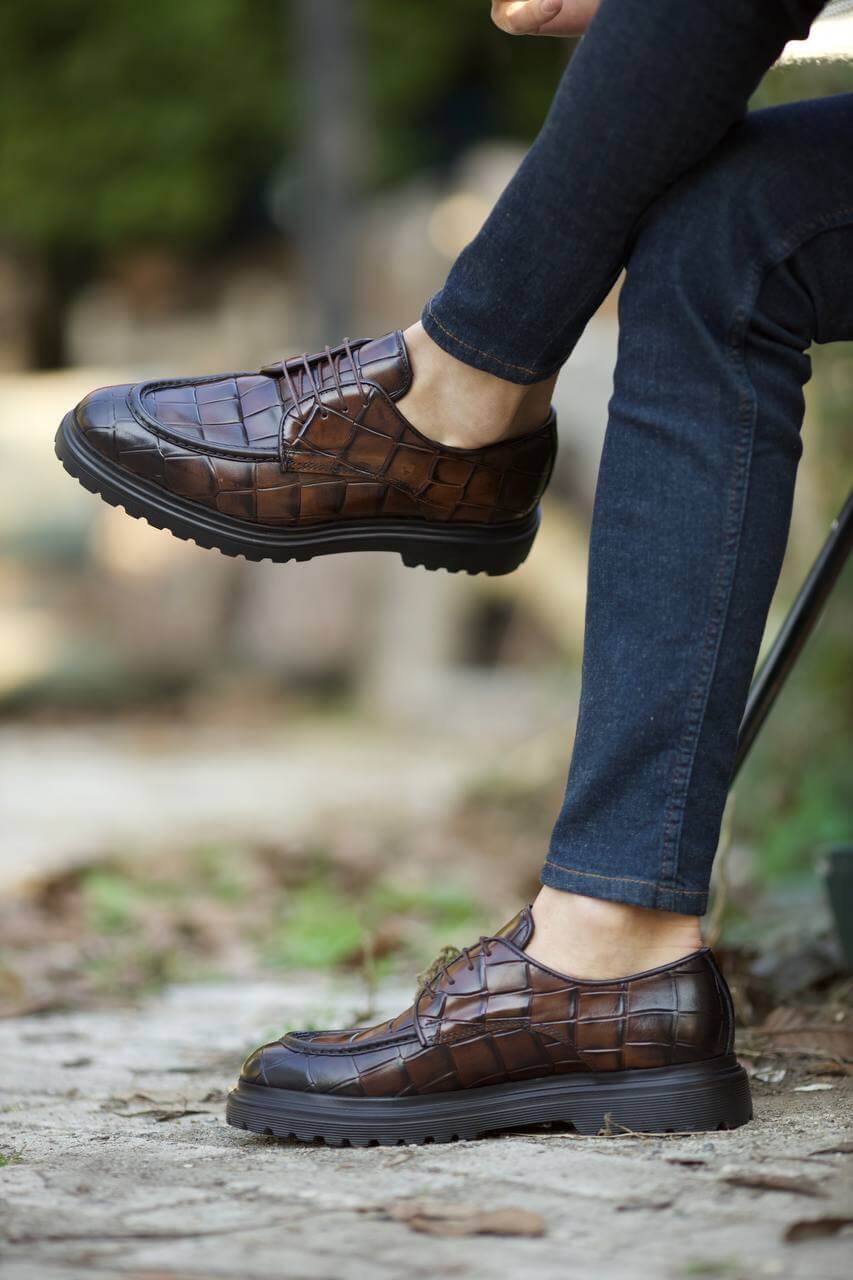 Close-up of a person wearing brown leather dress shoes with a textured pattern, stepping down on an old wooden staircase while dressed in dark denim jeans.