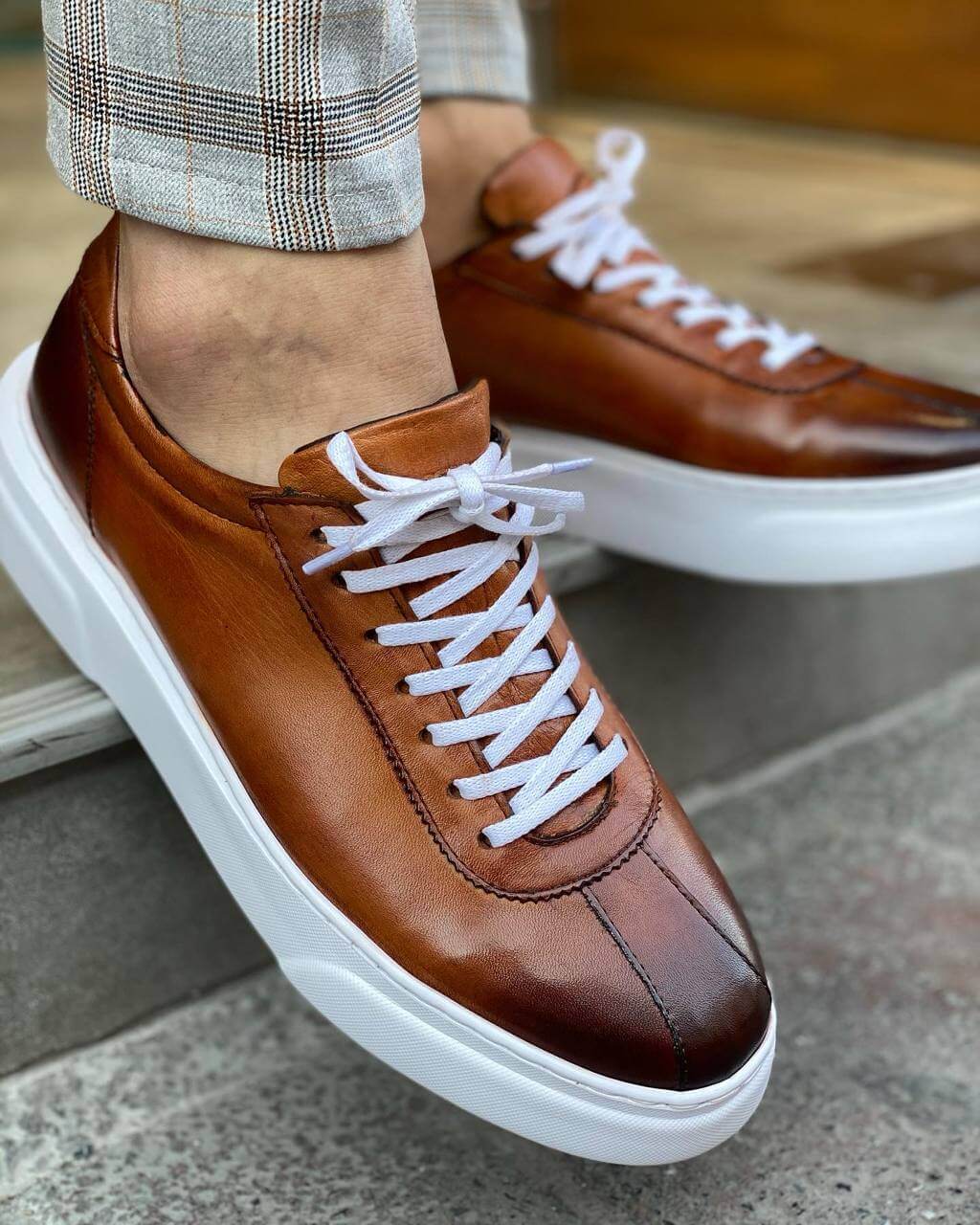 Close-up of a man wearing camel leather sneakers with white laces and a thick white sole, paired with stylish plaid trousers, standing on a stone step.