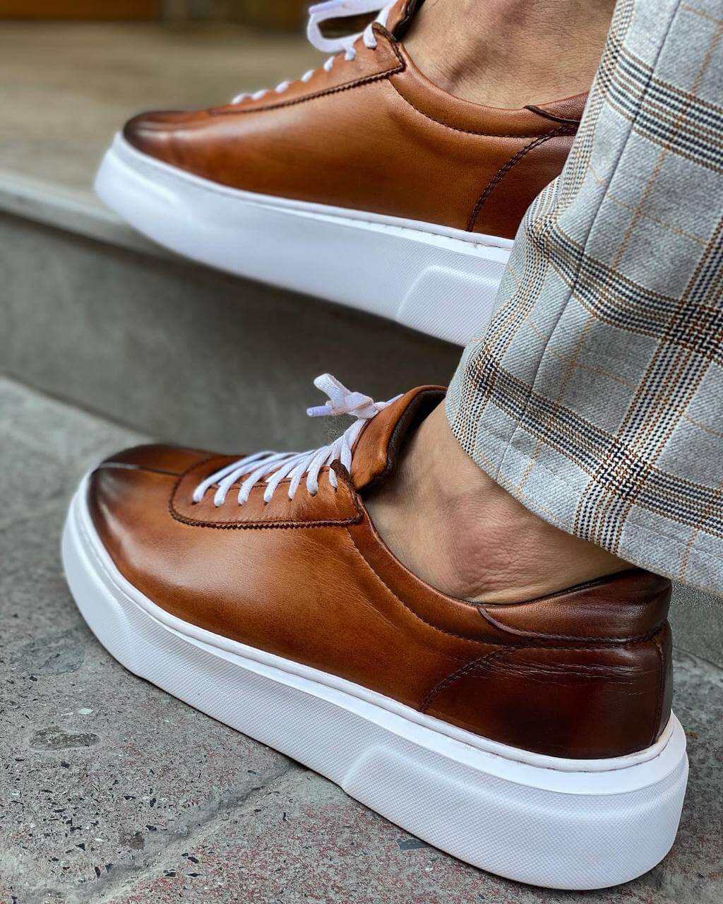 Close-up of a man wearing camel leather sneakers with white laces and a thick white sole, paired with stylish plaid trousers, standing on a stone step.