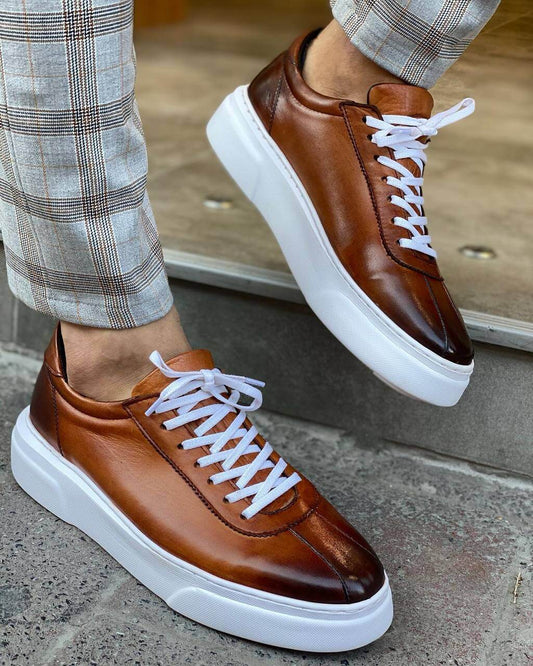 Close-up of a man wearing camel leather sneakers with white laces and a thick white sole, paired with stylish plaid trousers, standing on a stone step.