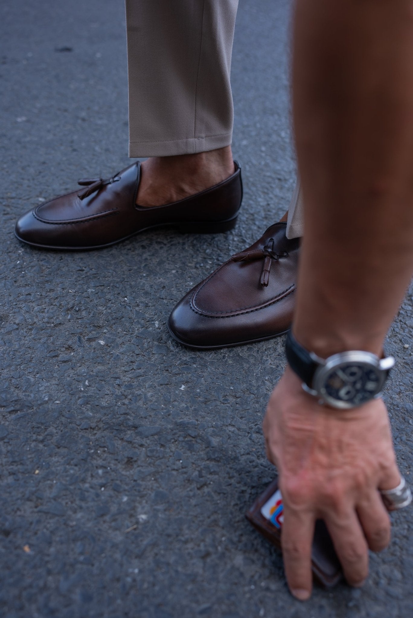 Close-up of a man wearing chestnut brown tassel loafers, paired with light beige trousers, standing on a textured asphalt surface.