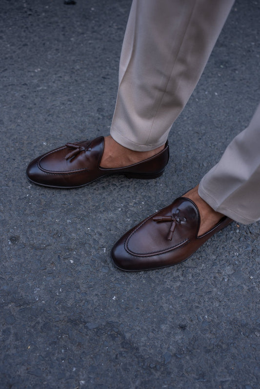 Close-up of a man wearing chestnut brown tassel loafers, paired with light beige trousers, standing on a textured asphalt surface.