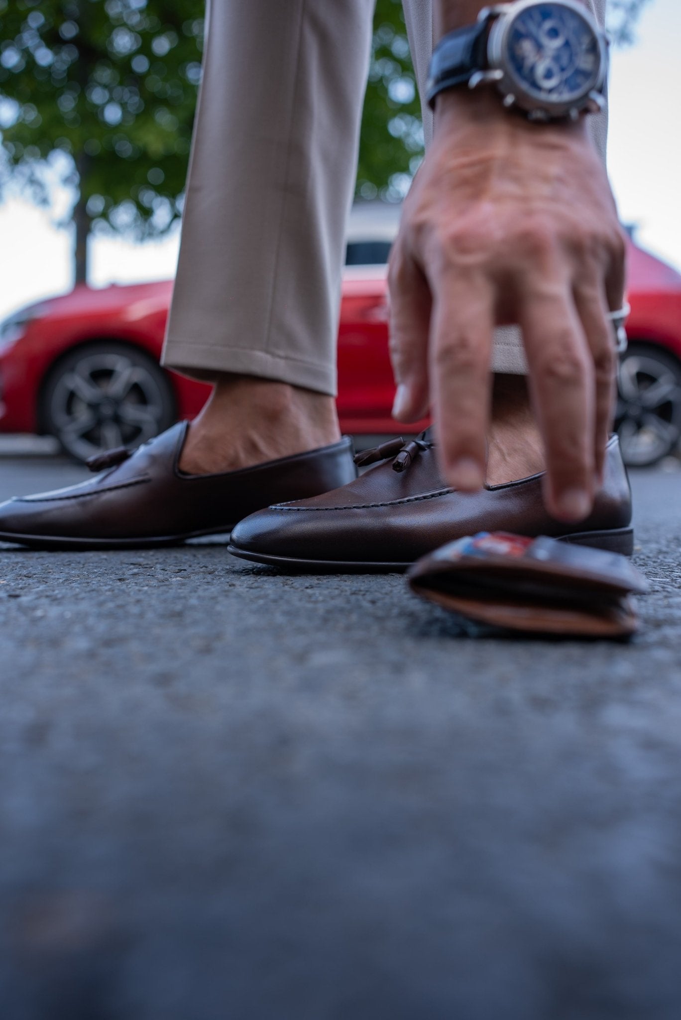 Close-up of a man wearing chestnut brown tassel loafers, paired with light beige trousers, standing on a textured asphalt surface.