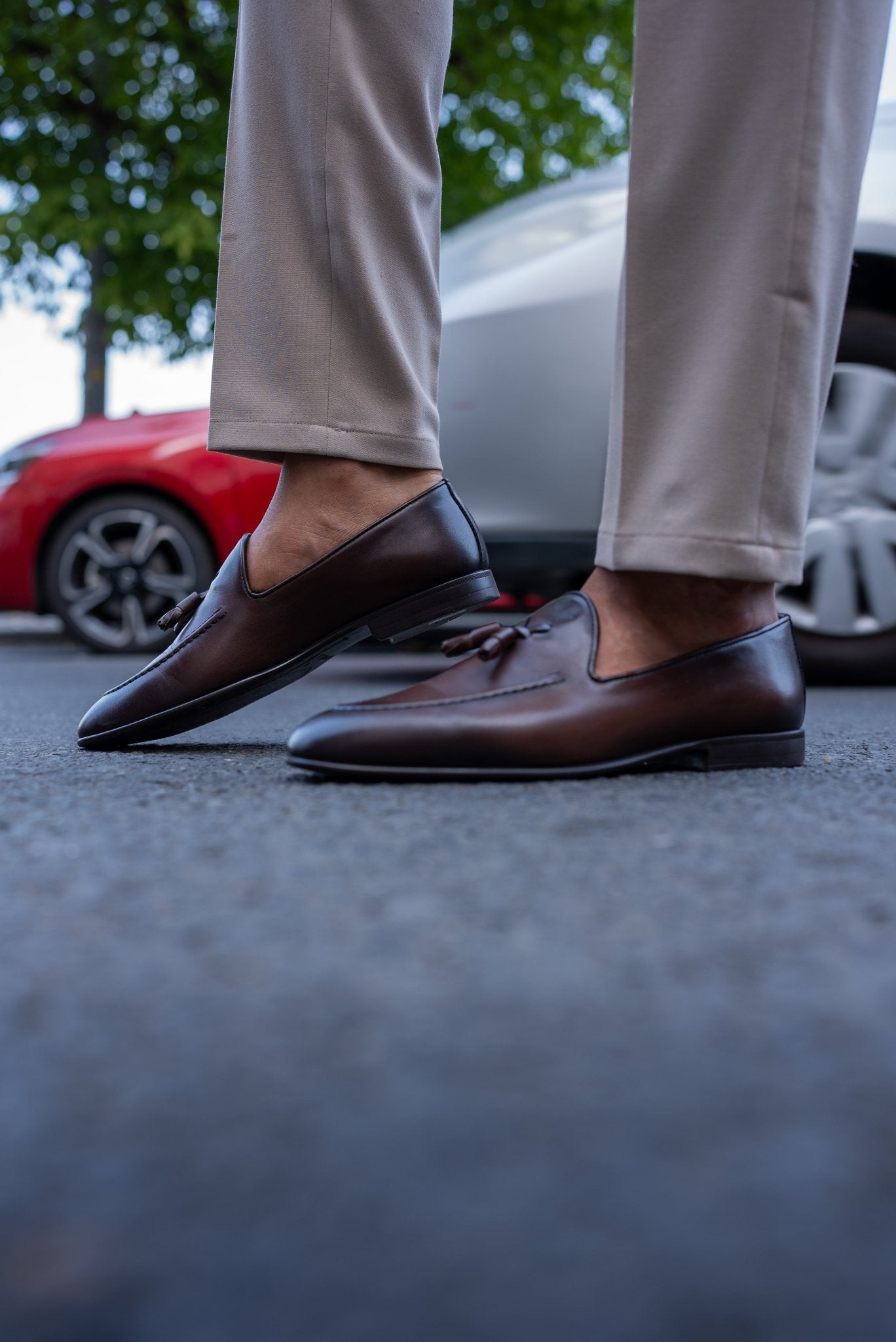 Close-up of a man wearing chestnut brown tassel loafers, paired with light beige trousers, standing on a textured asphalt surface.