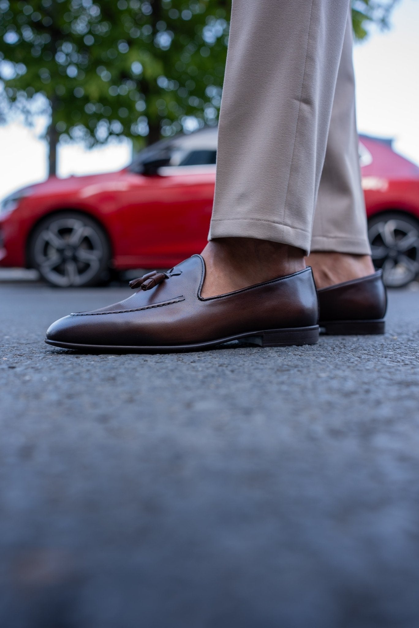 Close-up of a man wearing chestnut brown tassel loafers, paired with light beige trousers, standing on a textured asphalt surface.