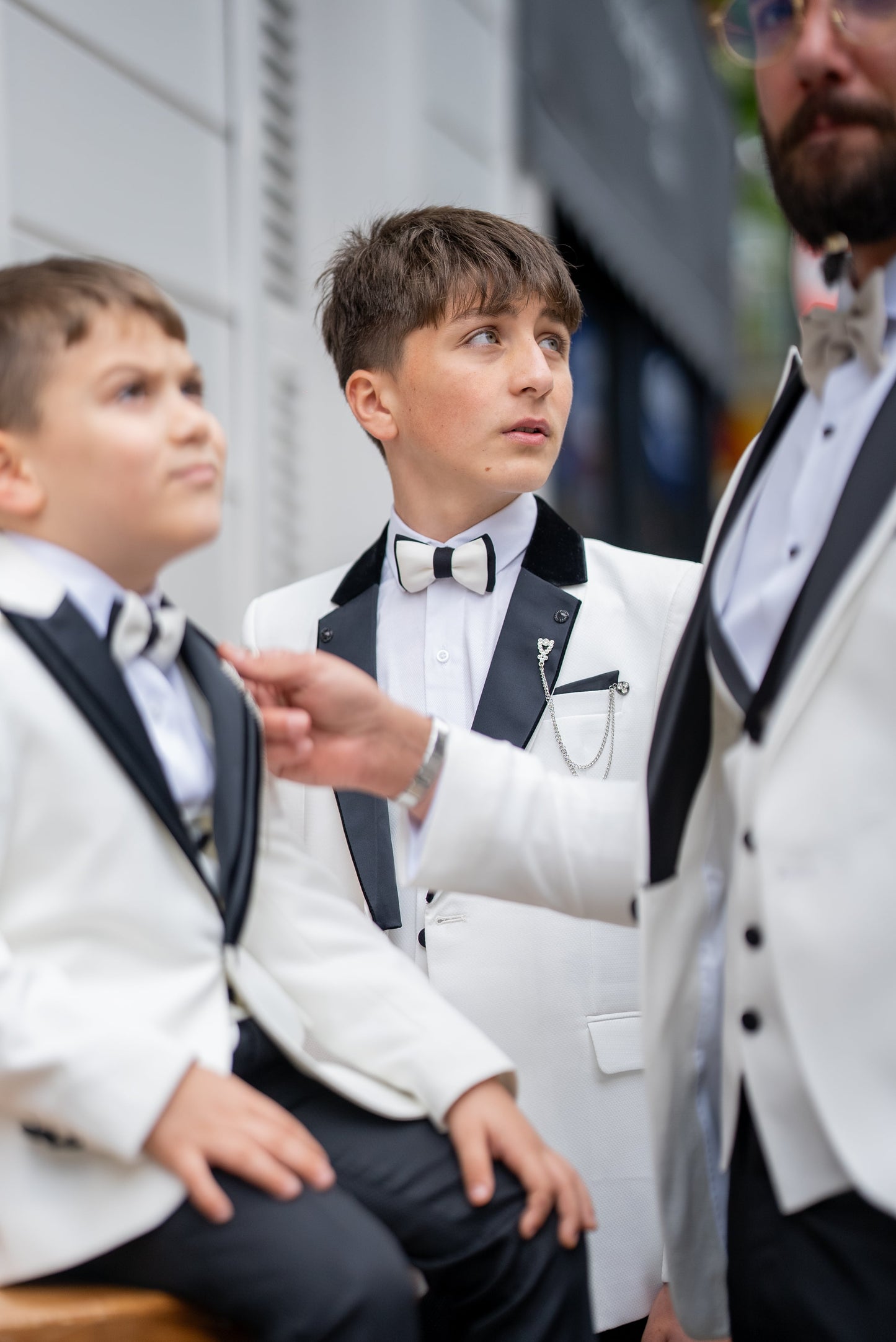 Elegant group shot of a father and his two sons dressed in matching white tuxedos with black accents, standing confidently in front of a stylish wooden door, ready for a formal event.