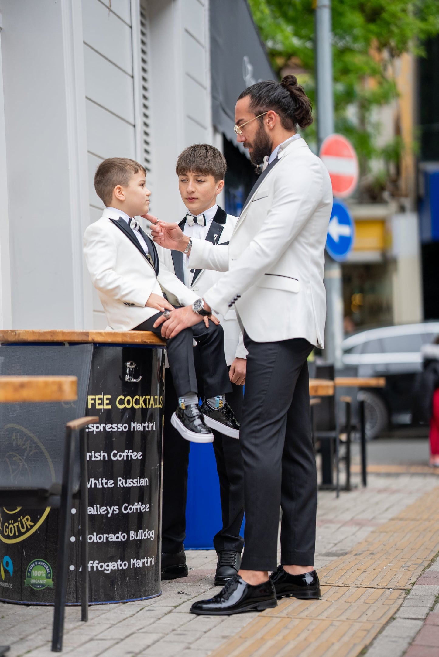 Elegant group shot of a father and his two sons dressed in matching white tuxedos with black accents, standing confidently in front of a stylish wooden door, ready for a formal event.