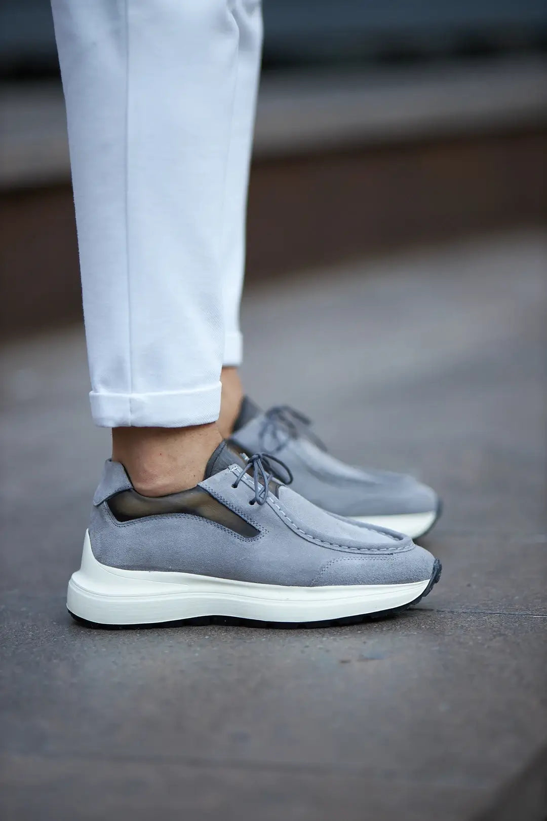 A stylish pair of gray suede smart casual shoes, designed by HolloMen, worn with white pants on a sidewalk.