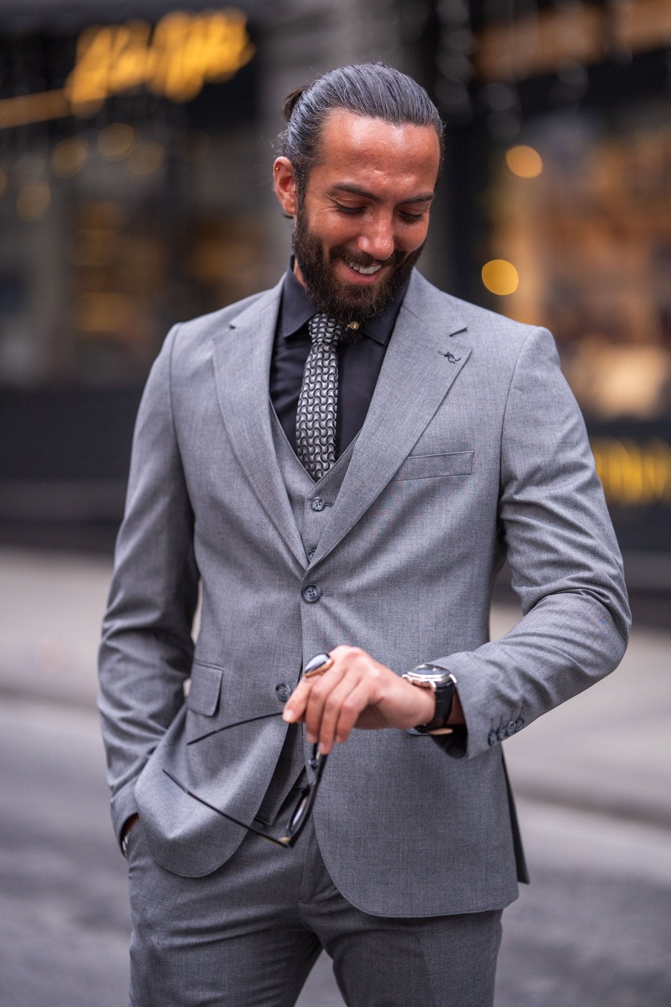 Confident man in a slim-fit gray suit with a black shirt and patterned tie, smiling while holding sunglasses, standing on an urban street with a blurred background.