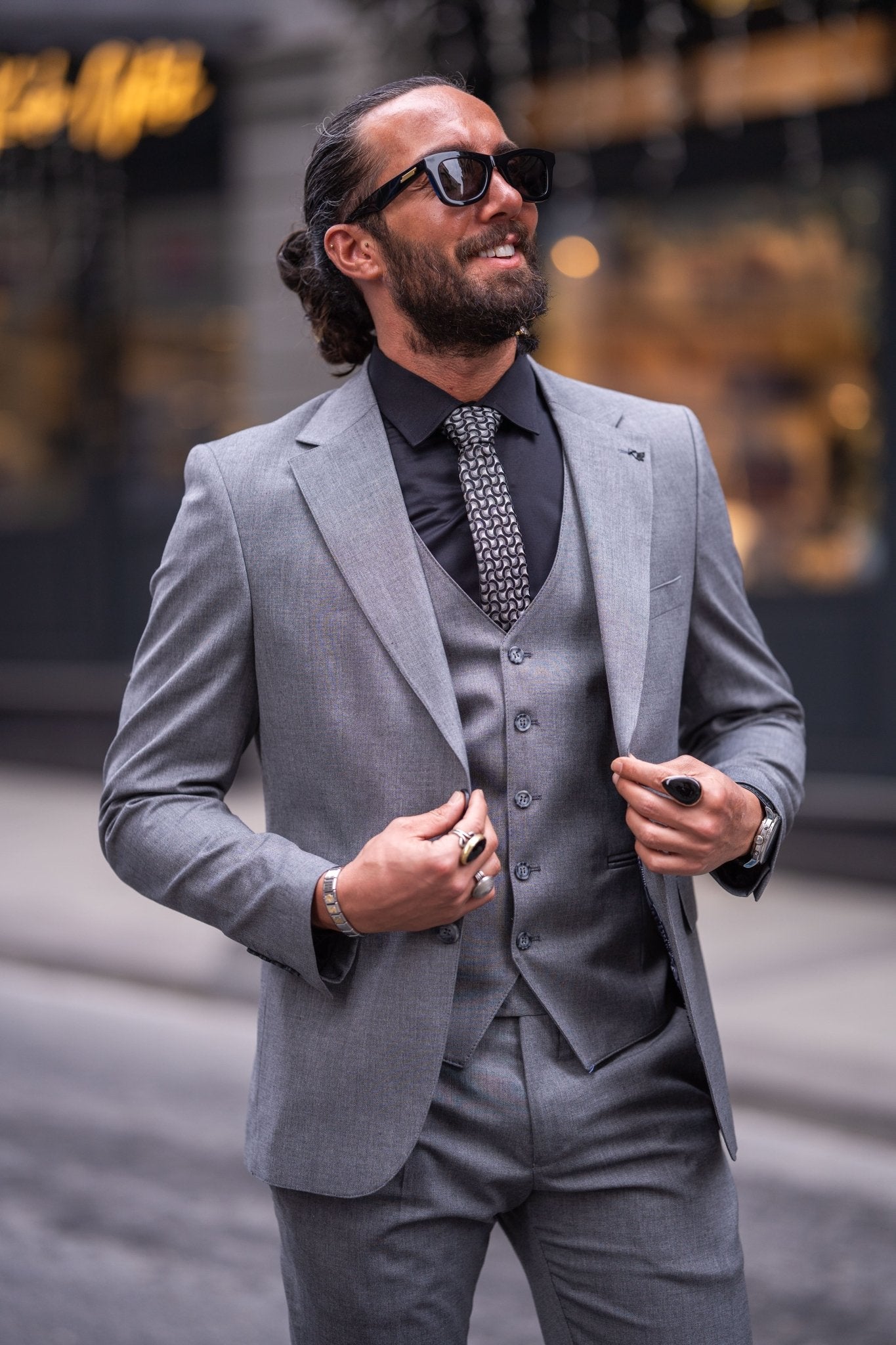 Confident man in a slim-fit gray suit with a black shirt and patterned tie, smiling while holding sunglasses, standing on an urban street with a blurred background.