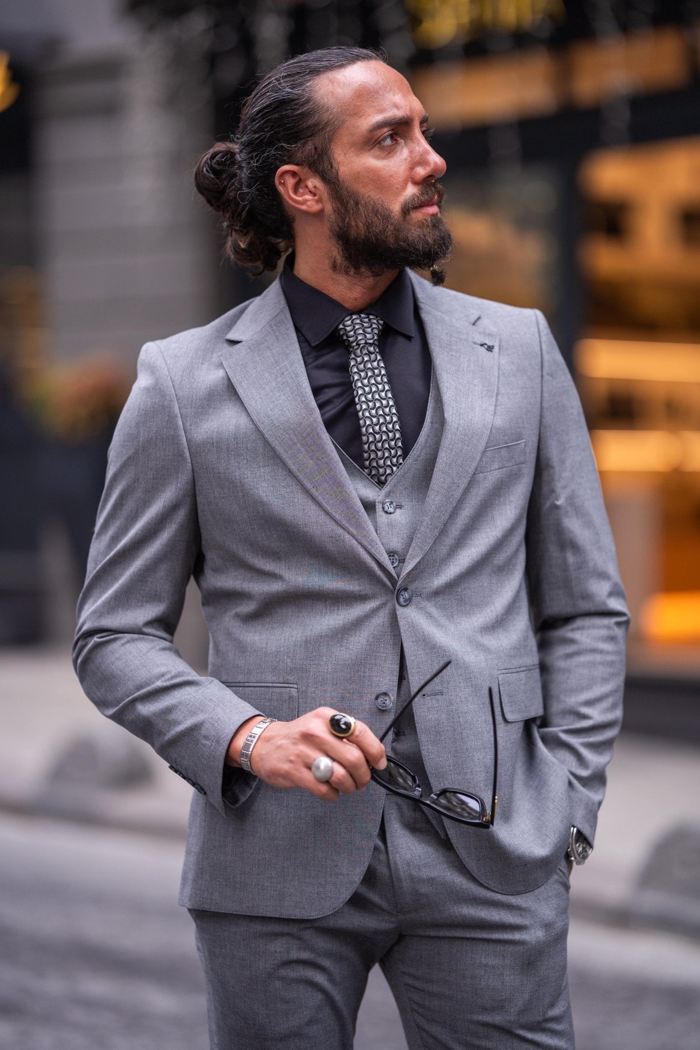 Confident man in a slim-fit gray suit with a black shirt and patterned tie, smiling while holding sunglasses, standing on an urban street with a blurred background.