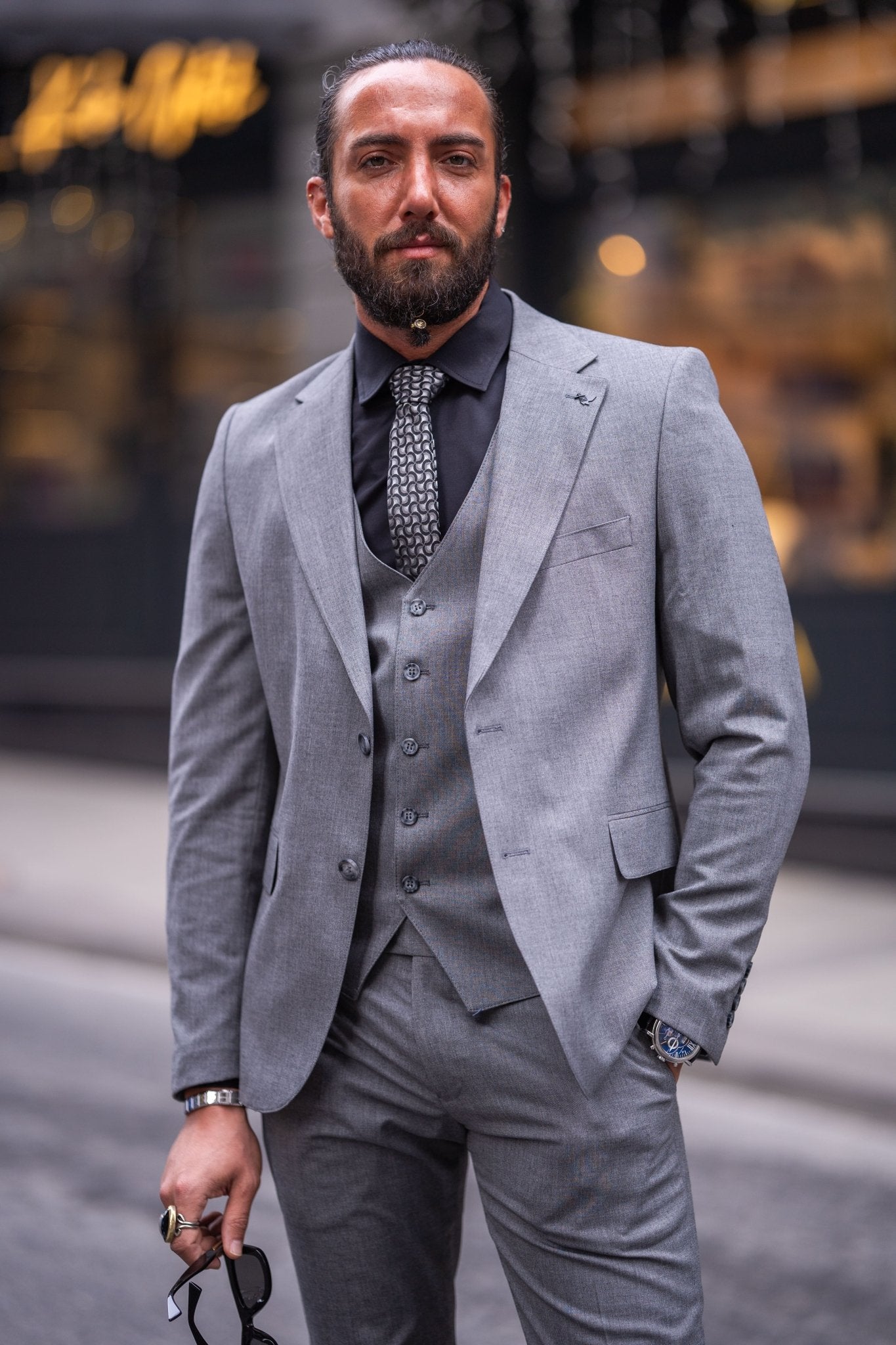 Confident man in a slim-fit gray suit with a black shirt and patterned tie, smiling while holding sunglasses, standing on an urban street with a blurred background.