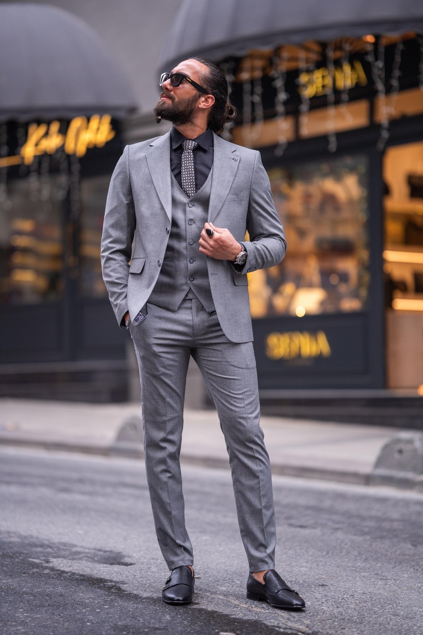 Confident man in a slim-fit gray suit with a black shirt and patterned tie, smiling while holding sunglasses, standing on an urban street with a blurred background.