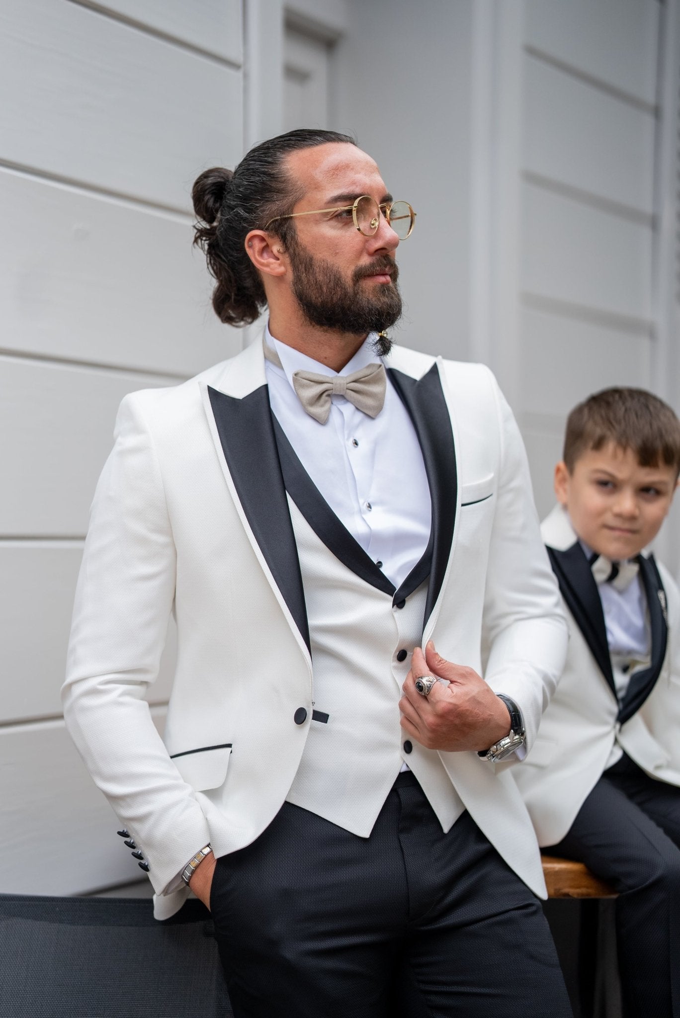 Elegant group shot of a father and his two sons dressed in matching white tuxedos with black accents, standing confidently in front of a stylish wooden door, ready for a formal event.