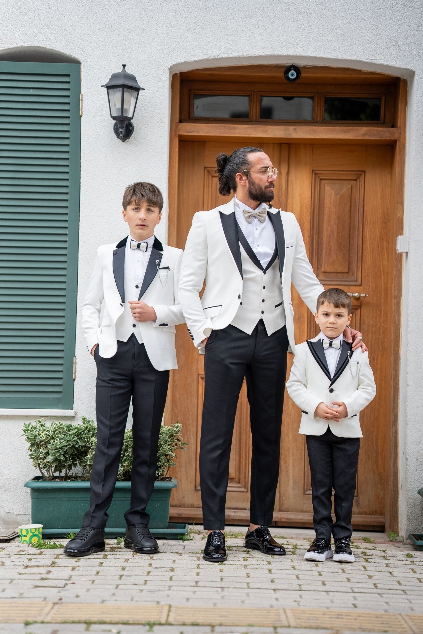 Elegant group shot of a father and his two sons dressed in matching white tuxedos with black accents, standing confidently in front of a stylish wooden door, ready for a formal event.