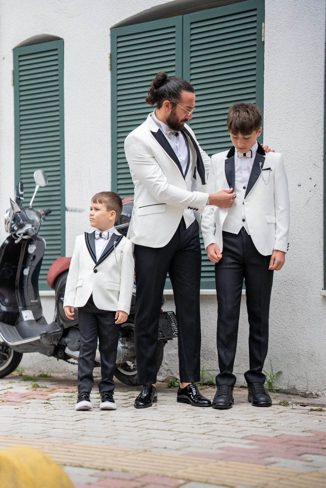 Elegant group shot of a father and his two sons dressed in matching white tuxedos with black accents, standing confidently in front of a stylish wooden door, ready for a formal event.