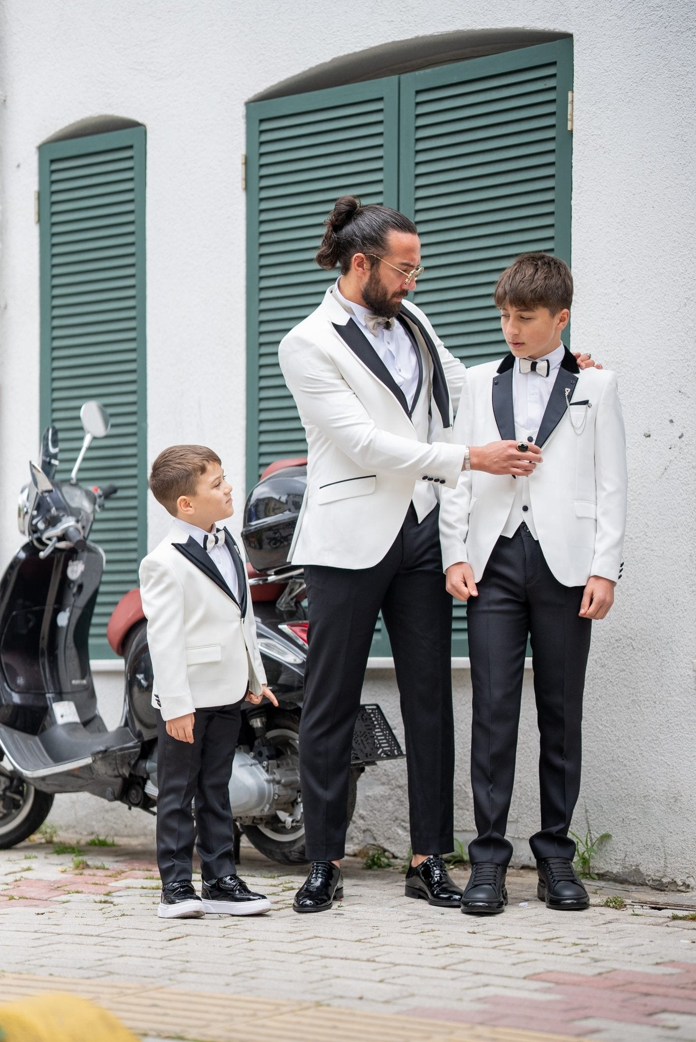 Elegant group shot of a father and his two sons dressed in matching white tuxedos with black accents, standing confidently in front of a stylish wooden door, ready for a formal event.