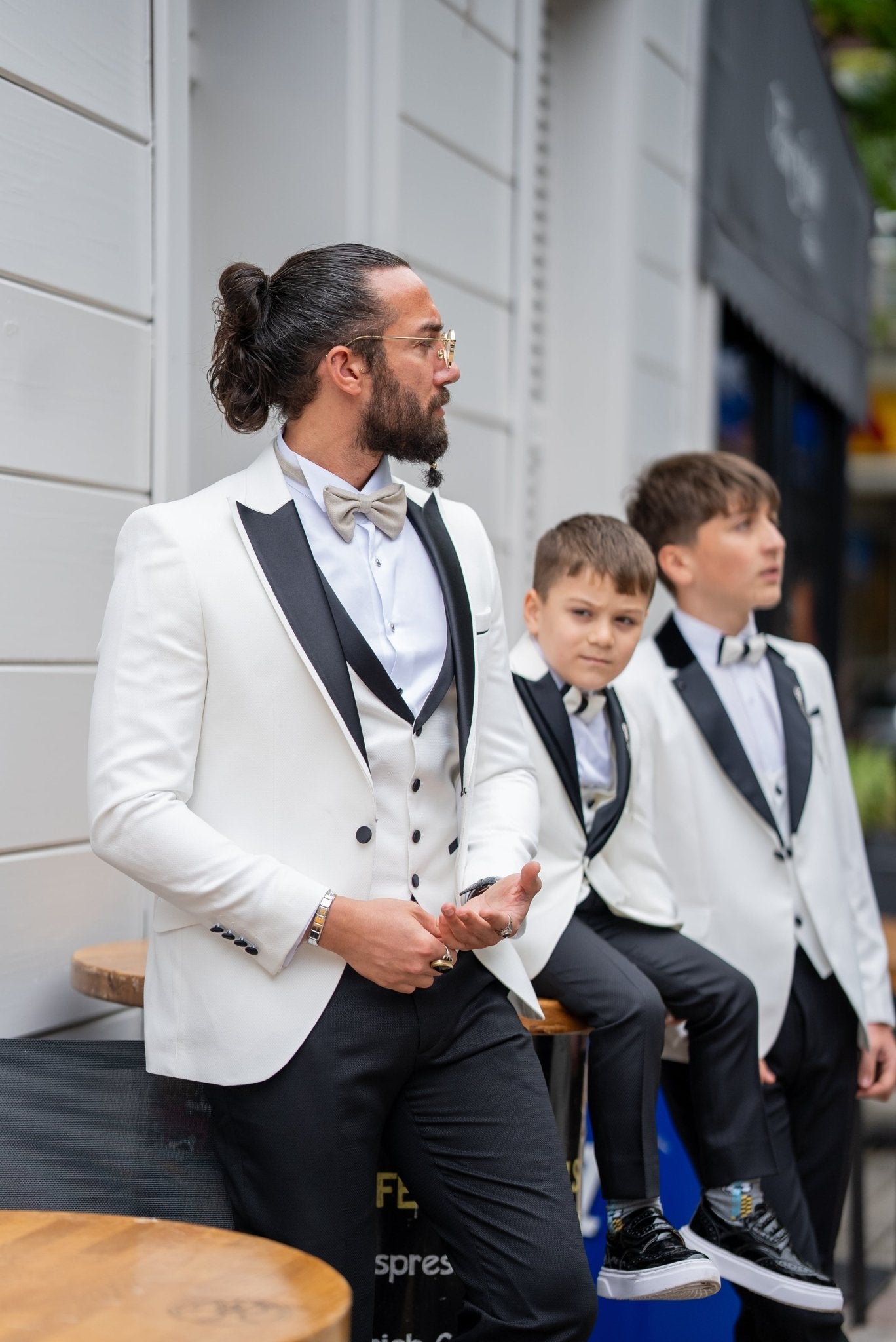 Elegant group shot of a father and his two sons dressed in matching white tuxedos with black accents, standing confidently in front of a stylish wooden door, ready for a formal event.