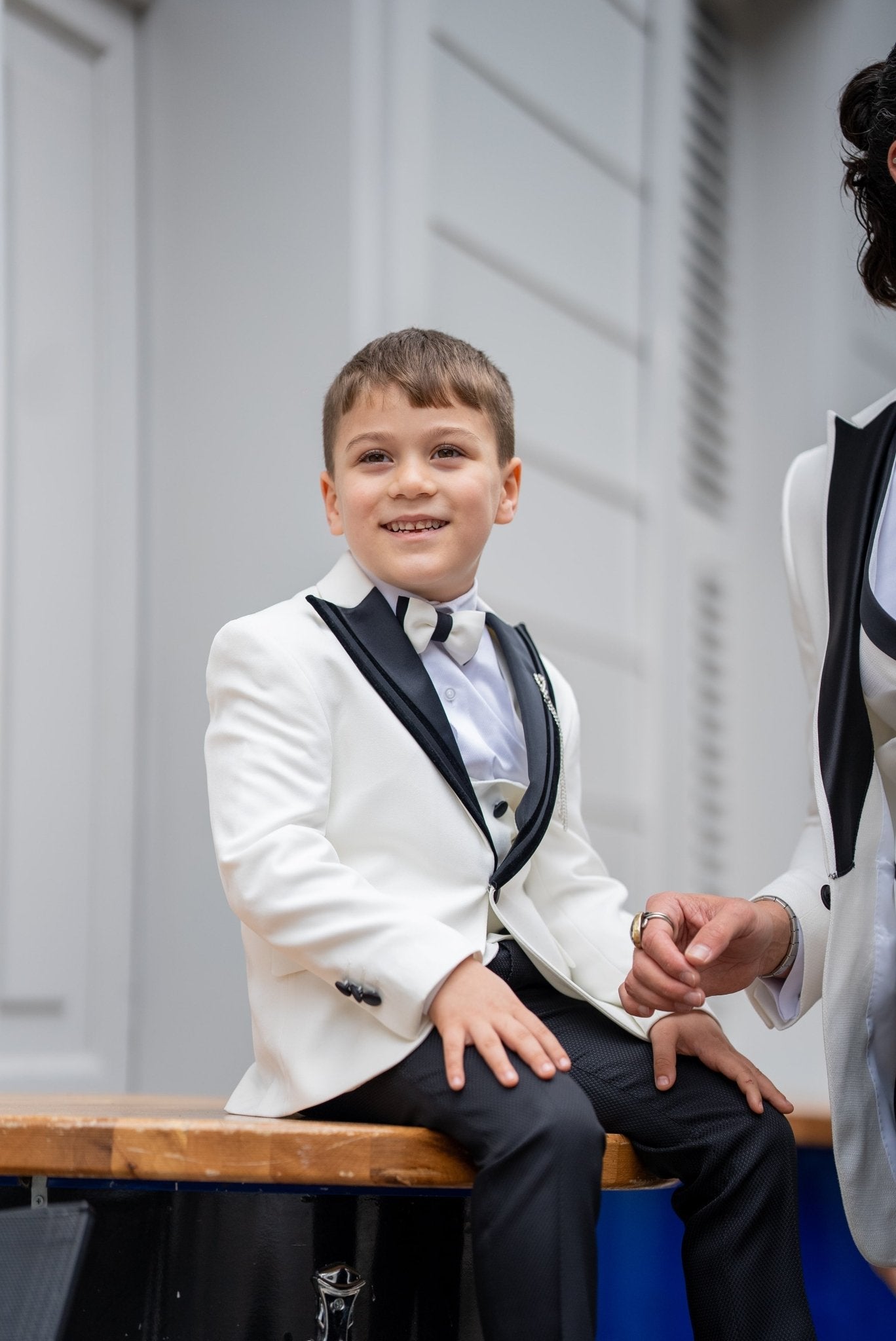 Elegant group shot of a father and his two sons dressed in matching white tuxedos with black accents, standing confidently in front of a stylish wooden door, ready for a formal event.