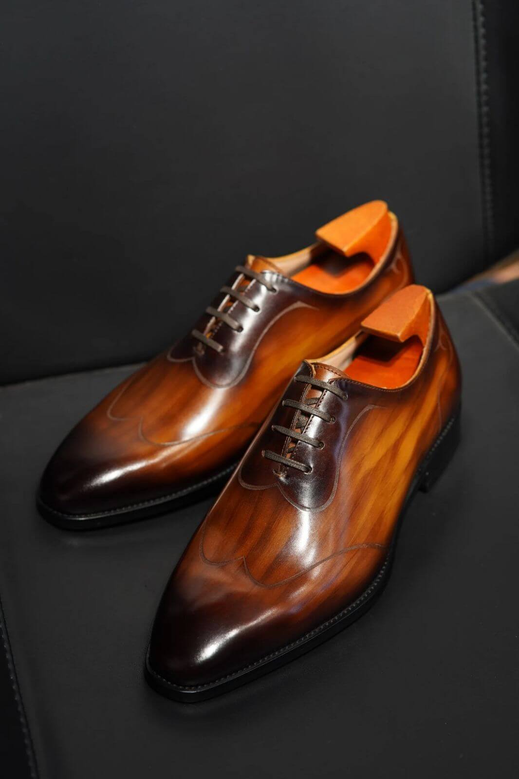 Close-up of a man wearing elegant amber brown patina Oxford shoes paired with black socks and pinstripe trousers, standing on polished wooden flooring