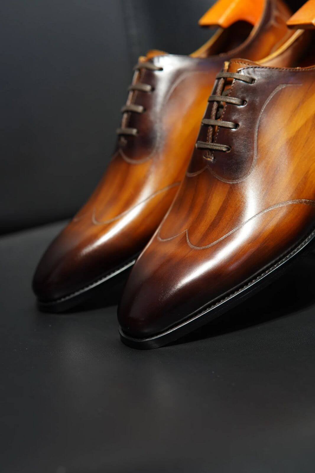 Close-up of a man wearing elegant amber brown patina Oxford shoes paired with black socks and pinstripe trousers, standing on polished wooden flooring