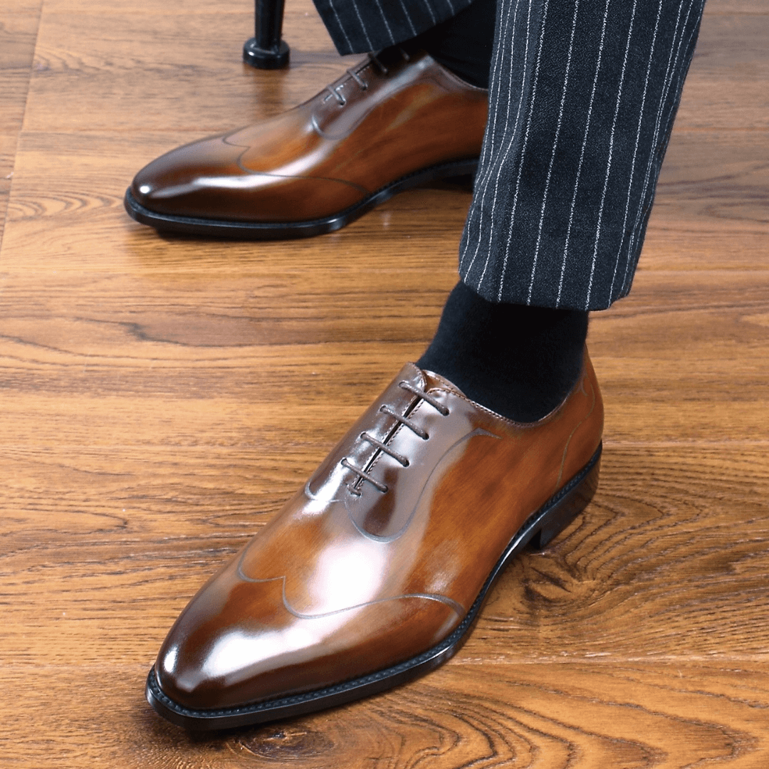 Close-up of a man wearing elegant amber brown patina Oxford shoes paired with black socks and pinstripe trousers, standing on polished wooden flooring