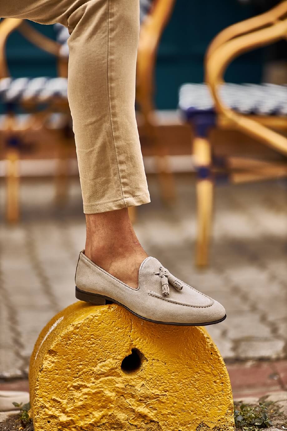 Man wearing beige suede tassel loafers paired with light khaki trousers, sitting outdoors on a patterned stone pavement.