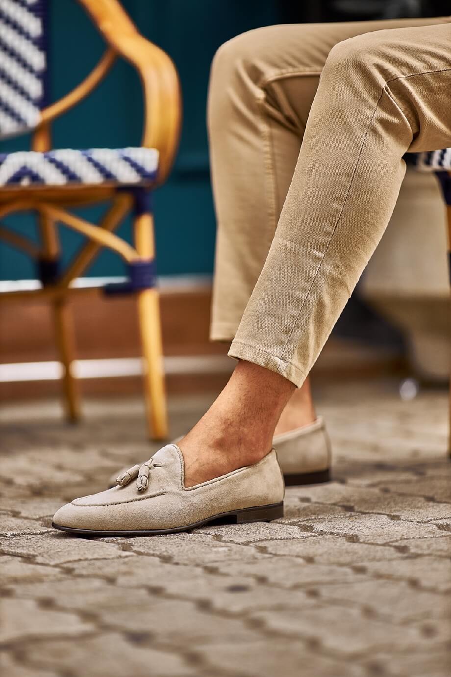 Man wearing beige suede tassel loafers paired with light khaki trousers, sitting outdoors on a patterned stone pavement.