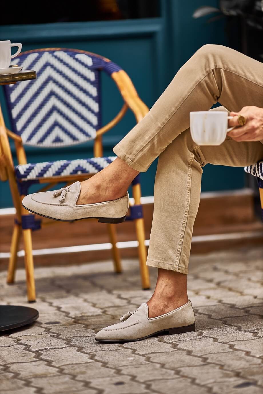 Man wearing beige suede tassel loafers paired with light khaki trousers, sitting outdoors on a patterned stone pavement.