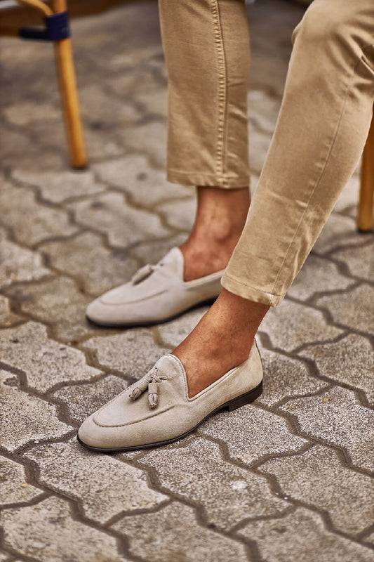 Man wearing beige suede tassel loafers paired with light khaki trousers, sitting outdoors on a patterned stone pavement.