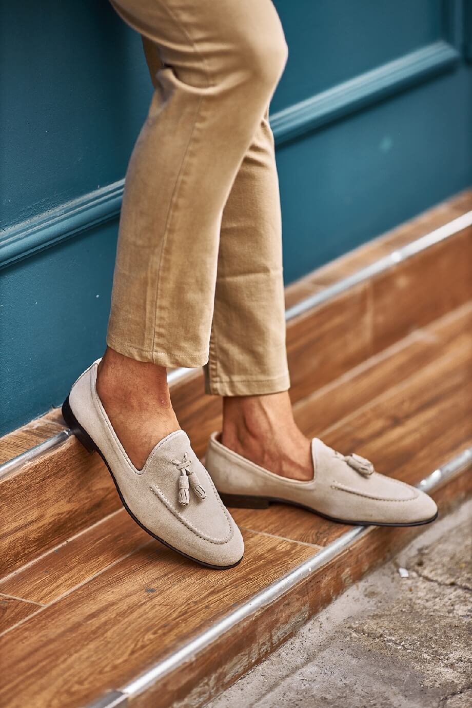 Man wearing beige suede tassel loafers paired with light khaki trousers, sitting outdoors on a patterned stone pavement.