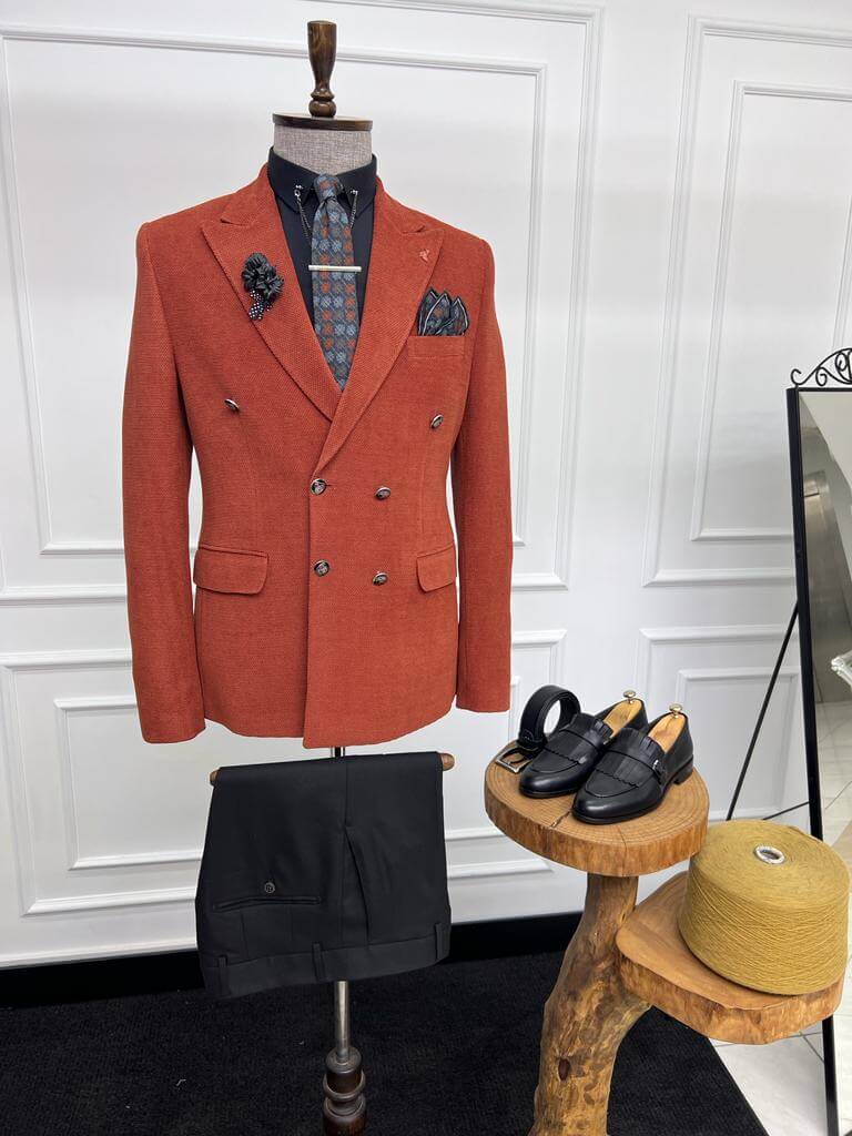 Stylish brick-red double-breasted blazer displayed on a mannequin, paired with a patterned tie, black shirt, and matching pocket square, complete with a lapel pin, set against a white paneled background.