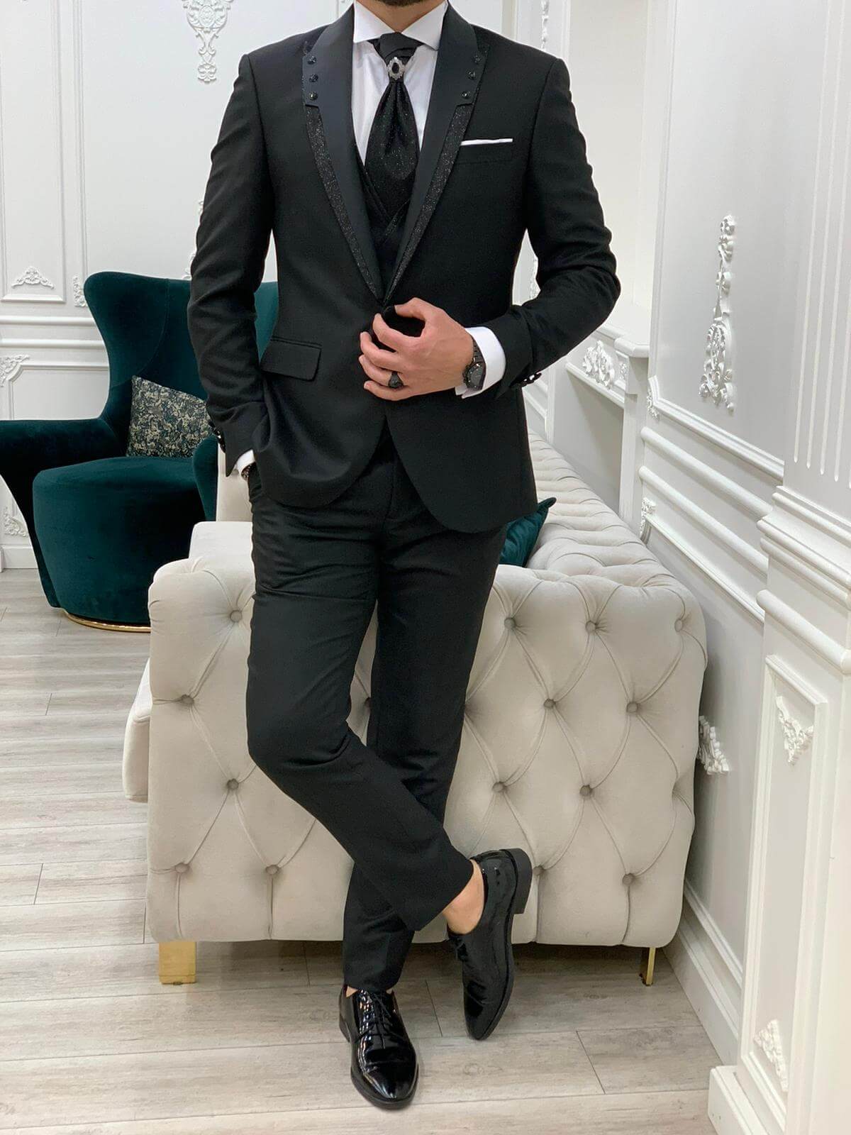 Man in a sophisticated black tuxedo with satin lapels and subtle embellishments, paired with a black tie and polished black dress shoes, standing confidently in a luxurious, well-appointed room.